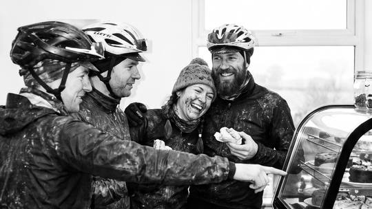 four mud-covered riders at a café counter, choosing cake