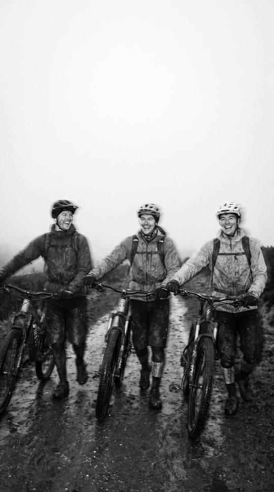 three riders walking their bikes back at dusk, arms round each other