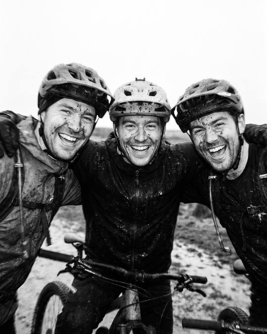 three mud-splattered riders, arms round each other, grinning at the camera
