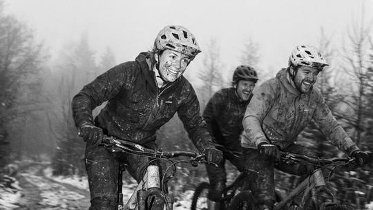 three riders grinning as they ride a muddy, snow-dusted forest track