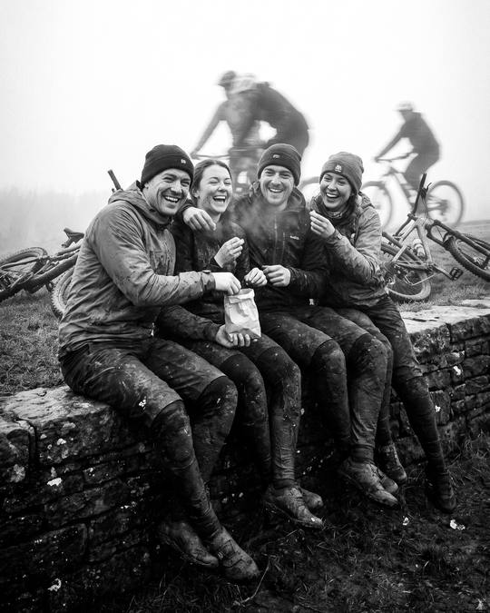 four riders sat on a stone wall sharing a bag of snacks, bikes in the grass behind